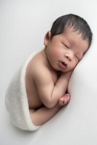 Newborn baby sleeping on their side, wrapped in a white blanket, against a soft white background.