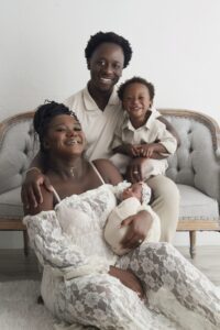 A family of four poses on a couch: a woman in a white lace dress holds a swaddled baby, with a man and a young boy in light clothing smiling beside her.