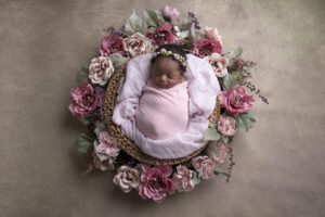 Newborn baby wrapped in a pink blanket, sleeping in a basket surrounded by pink and mauve artificial flowers, with a flower headband on her head.