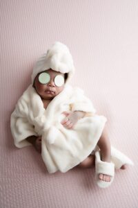 Baby wearing a white robe, matching towel, and slippers, lying on a pink surface with cucumber slices over their eyes, imitating a spa treatment.