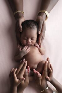 A newborn baby lies on a pink textured surface, gently cradled by several adult and child hands wearing gold jewelry.