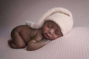 Newborn baby sleeping on a textured pink blanket, wearing a soft, light pink knit hat with a long tail.