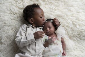 A young child in a beige shirt lies on a soft, white rug, gently kissing the forehead of a sleeping baby dressed in a white outfit.