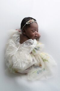 A newborn baby wearing a floral headband and a white butterfly wing costume sleeps on a white surface.