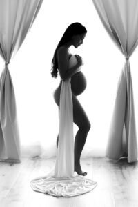 Black and white photo of a pregnant woman in profile, standing between curtains, draped in a white cloth with natural light in the background—an elegant example of the timeless style you can achieve with studio vs beach maternity photos Miami.