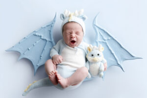 Baby dressed in a blue dragon costume, sitting with a matching dragon plush toy, yawning or crying against a light blue background.