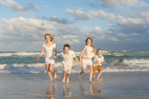 Four children wearing light-colored clothes run together on a sandy beach near the ocean, with waves and clouds in the background—perfect for capturing fall family photos in Miami.
