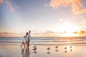 A family of three walks barefoot along the beach at sunset, enjoying fall family photos in Miami, with several seagulls on the wet sand and gentle waves in the background.