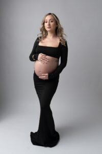 Pregnant woman in a long black dress stands on a plain background, holding her bare belly with both hands and looking at the camera—capturing the elegance of studio maternity photography, perfect for those considering studio vs beach maternity photos in Miami.