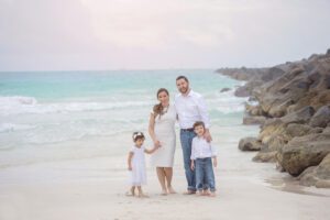Family of four stands barefoot on a sandy beach near rocks and ocean, wearing white and light-colored clothing, posing and smiling at the camera—capturing beautiful fall family photos in Miami that showcase their joy by the shoreline.