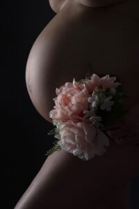 A close-up of a pregnant belly in profile, with a bouquet of pink and white flowers resting gently against the skin, set against a dark background—perfectly capturing the timeless elegance found in studio maternity photos Miami moms adore, as opposed to the sunlit beach style.