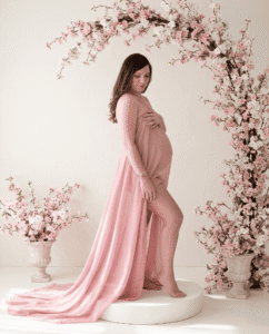 Pregnant woman in a flowing pink gown stands on a white platform, surrounded by pink cherry blossom branches in vases and an arch, capturing the elegance and timeless beauty often seen in studio vs beach maternity photos Miami.