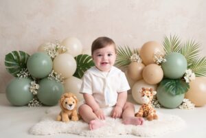 A baby sits on a white rug between a stuffed lion and giraffe, with green, tan, and white balloons and palm leaves in the background.
