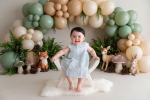 A smiling baby sits on a white chair in front of a balloon arch with forest animal decorations, mushrooms, and ferns.