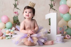 Baby in a crown and tutu sits next to a large number one, surrounded by balloons, flowers, cupcakes, and a cake, suggesting a first birthday celebration.