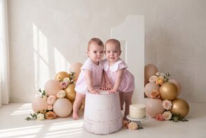 Two babies in matching pink outfits pose with a white wooden stool, surrounded by balloons, flowers, a cake, and a large number one decoration.