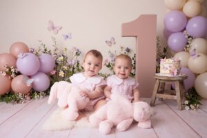 Two babies in matching outfits sit with stuffed bunnies in front of a large number one, surrounded by pastel balloons, flowers, and a cake on a stool.