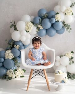 A baby in a blue floral dress sits on a white chair with blue and white balloons and flowers behind her, and a cake with blue and white decorations on a stand nearby.