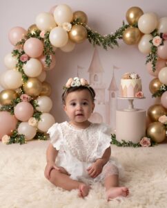 A toddler in a white dress and floral headband sits on a fluffy rug in front of a balloon arch and a cake, with a castle backdrop.