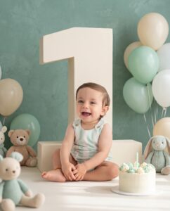 Smiling baby sitting in front of a large number one, surrounded by stuffed animals, balloons, and a birthday cake with one candle.