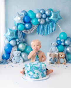 Baby sits in front of a blue and white cake, surrounded by blue balloons and stuffed animals, wearing a blue tie.