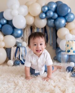 A smiling toddler in a white shirt and denim shorts crawls on a fluffy rug, with blue and white balloons and party decorations in the background.