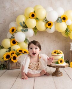 A baby in a white outfit sits on the floor beside a sunflower cake, with a sunflower and balloon arch decoration in the background.