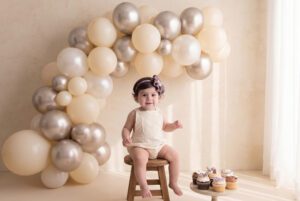 A toddler sits on a wooden stool in front of a balloon arch, with cupcakes on a small table nearby, in a softly lit room.