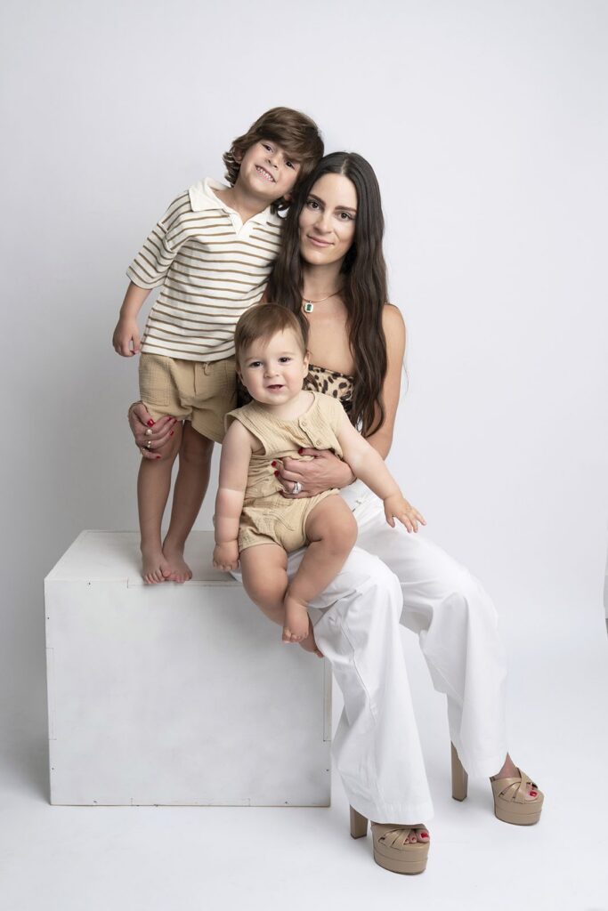 A woman sits on a white cube holding a baby, with an older child standing beside her. All three wear neutral-colored outfits against a white background, capturing the warmth and togetherness that makes the best time for family photos in Miami unforgettable.