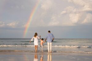 A woman, man, and child walk toward the ocean holding hands, with a rainbow visible in the cloudy sky over the water—capturing a magical moment that highlights the best time for family photos in Miami.